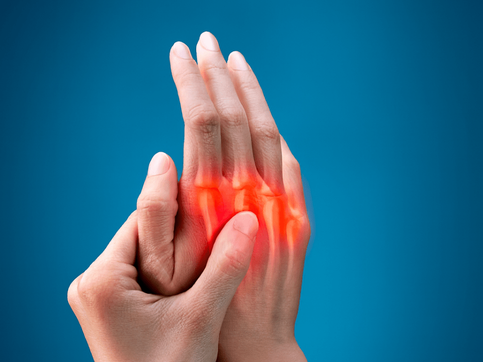 Imagem ilustrativa de uma pessoa segurando a mão com destaque em vermelho nas articulações, representando dor ou inflamação nas juntas.