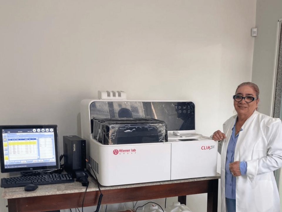Biomédica ao lado do equipamento Clia 900 da Wiener lab., instalado no Hospital e Maternidade Santo Antônio, em Itaituba (PA), demonstrando o uso de tecnologia de quimioluminescência para agilizar diagnósticos laboratoriais na região Norte do Brasil.