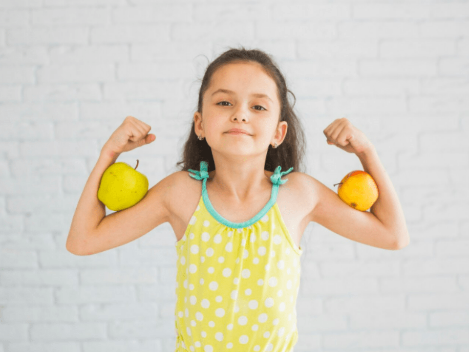 Criança sorridente com expressão de força, segurando uma maçã e uma pera nos braços, simbolizando saúde, nutrição e energia infantil.