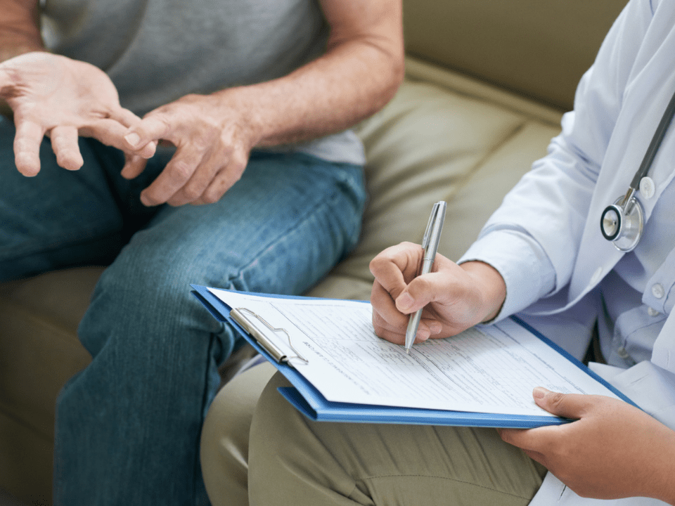 Paciente conversando com profissional de saúde sentado, enquanto o médico faz anotações em uma prancheta durante consulta, ambos com foco na comunicação e registro de informações clínicas.