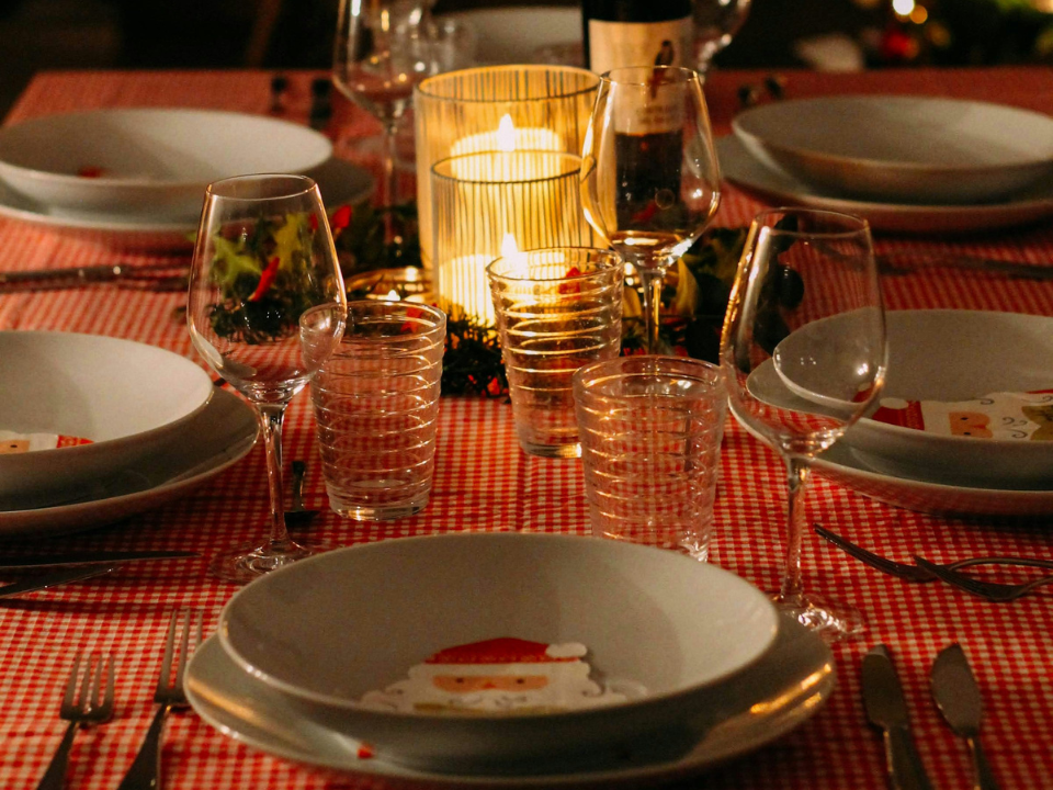 Mesa posta para ceia de Natal, com toalha xadrez vermelha e branca, pratos com detalhe do Papai Noel, taças, copos, talheres organizados e velas acesas ao centro, criando um ambiente acolhedor e festivo.