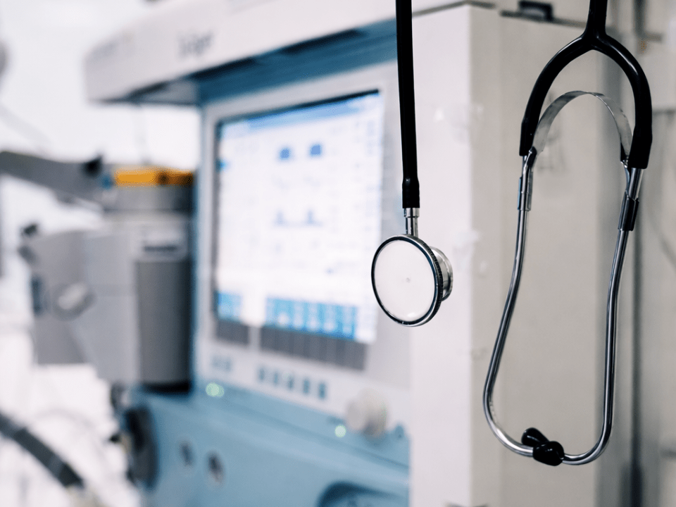 Estetoscópio pendurado em equipamento hospitalar, com monitor médico e dispositivos de suporte à vida ao fundo, em ambiente clínico controlado.