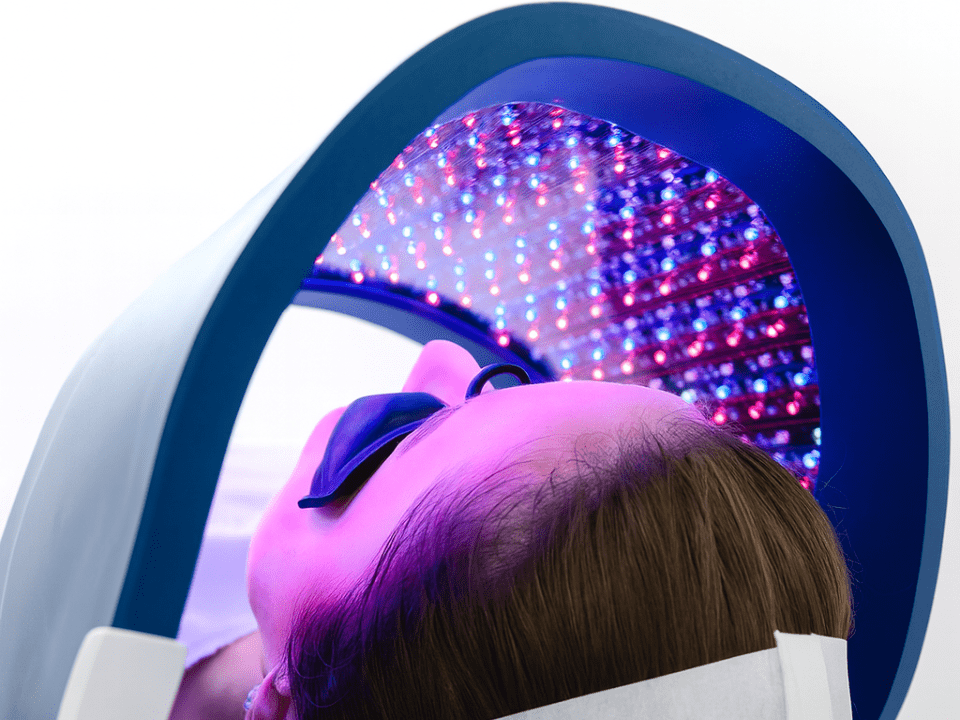 Mulher deitada em maca de clínica estética durante sessão de fototerapia com painel de LED azul e vermelho para tratamento de acne no rosto.