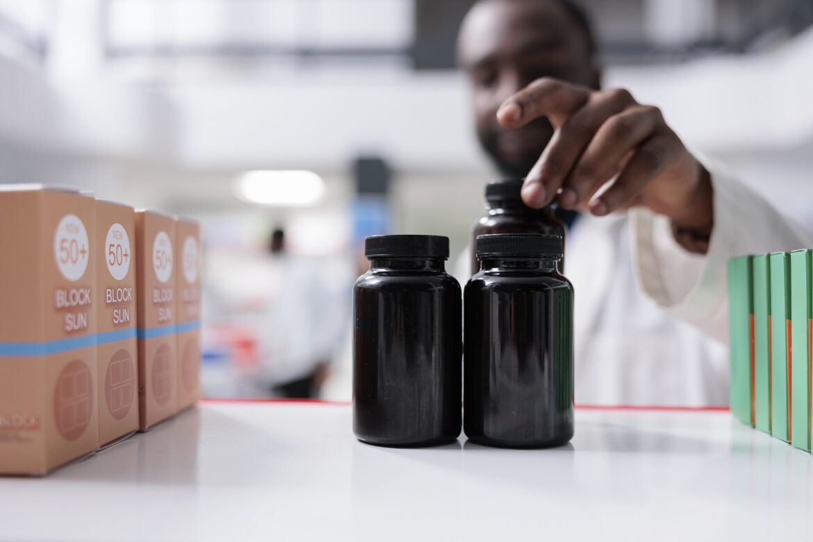 Profissional em ambiente de laboratório organiza frascos escuros de produtos cosméticos sobre bancada, com embalagens de protetor solar 50+ ao fundo, representando controle, envase e qualidade na indústria cosmética e farmacêutica.