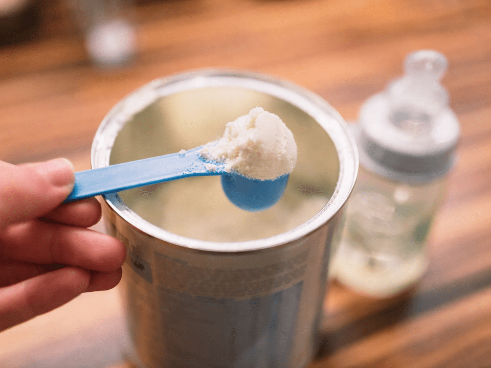 Fórmula infantil em pó sendo medida com colher dosadora, imagem ilustrativa de proibição de fórmulas infantis por risco de contaminação e alerta de vigilância sanitária.