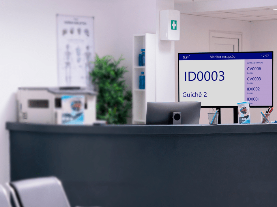 Recepção de centro de medicina diagnóstica com balcão de atendimento e monitor exibindo chamada de senha, ilustrando sistema digital de gestão de atendimento ao paciente.
