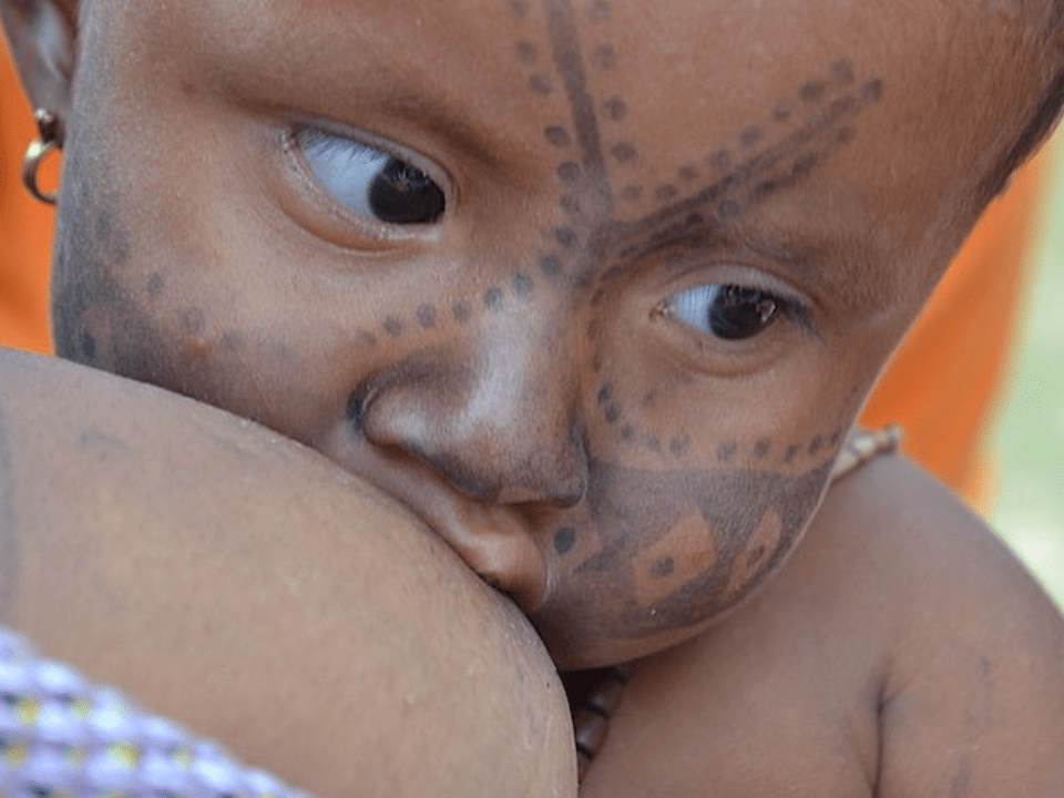 Bebê indígena em close, com pintura tradicional no rosto, sendo amamentado, representando cuidado materno e saúde na primeira infância.