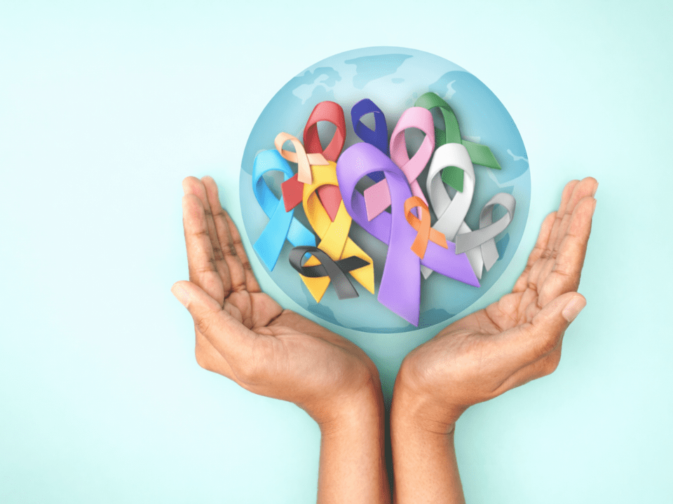 Mãos humanas sustentando um globo translúcido com laços coloridos de conscientização, simbolizando o Dia Mundial do Câncer, tendências em 2026 para prevenção, cuidado e impacto global da doença.
