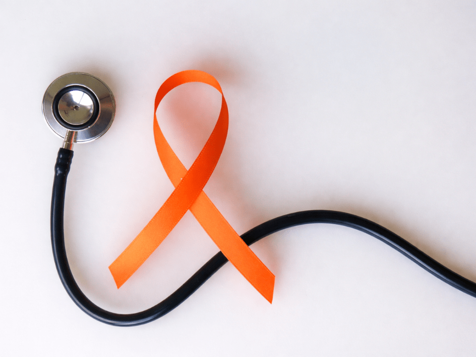 Estetoscópio disposto sobre fundo claro ao lado de um laço laranja, símbolo da campanha Fevereiro Laranja de conscientização sobre a leucemia e a doação de medula óssea.