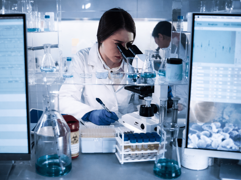 Pesquisadora em laboratório analisa amostras biológicas ao microscópio, com apoio de dados digitais, em um ambiente de pesquisa biomédica avançada.