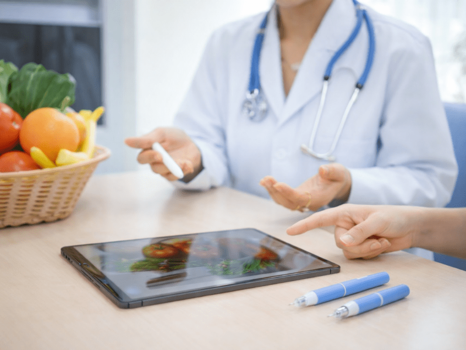 Médico orienta paciente durante consulta sobre uso de canetas emagrecedoras, com apoio de avaliação nutricional, alimentos frescos sobre a mesa e tablet com imagens de dieta saudável.