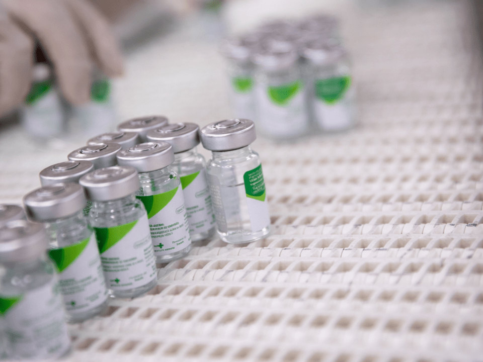 Frascos de vacina organizados em linha de produção farmacêutica, com mãos enluvadas manipulando os imunizantes em ambiente industrial esterilizado.