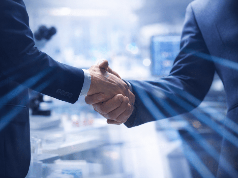 Aperto de mãos entre dois executivos em primeiro plano, com laboratório de biotecnologia desfocado ao fundo, simbolizando parceria estratégica no setor de diagnóstico em patologia.