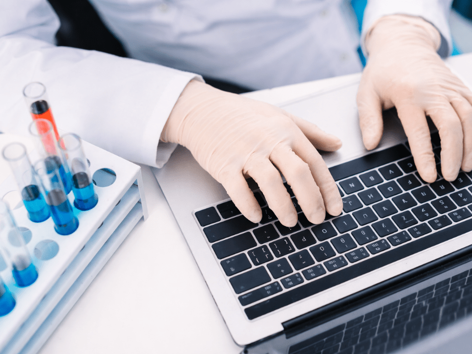 Profissional de laboratório com luvas manipulando laptop ao lado de tubos de ensaio com soluções coloridas, representando análise de dados e controle de qualidade laboratorial.