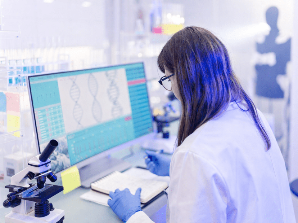 Cientista em laboratório analisa dados genéticos em computador, com microscópio e tubos ao fundo, em ambiente de diagnóstico molecular moderno.