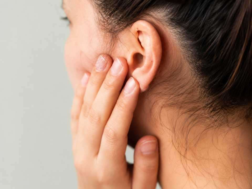 ￼Mulher toca suavemente a região da orelha em imagem de close-up, representando sintomas relacionados a zumbido e perda auditiva. A fotografia ilustra sinais precoces de alterações auditivas que podem impactar a comunicação, a qualidade de vida e a saúde auditiva.