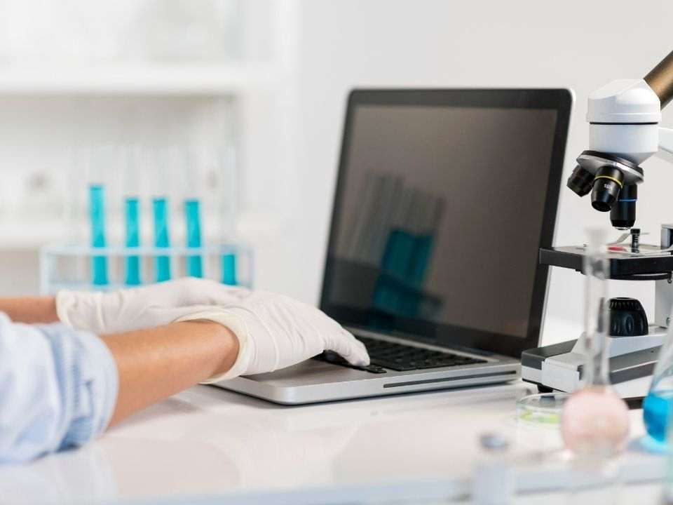Profissional de laboratório utiliza notebook ao lado de microscópio e tubos de ensaio.